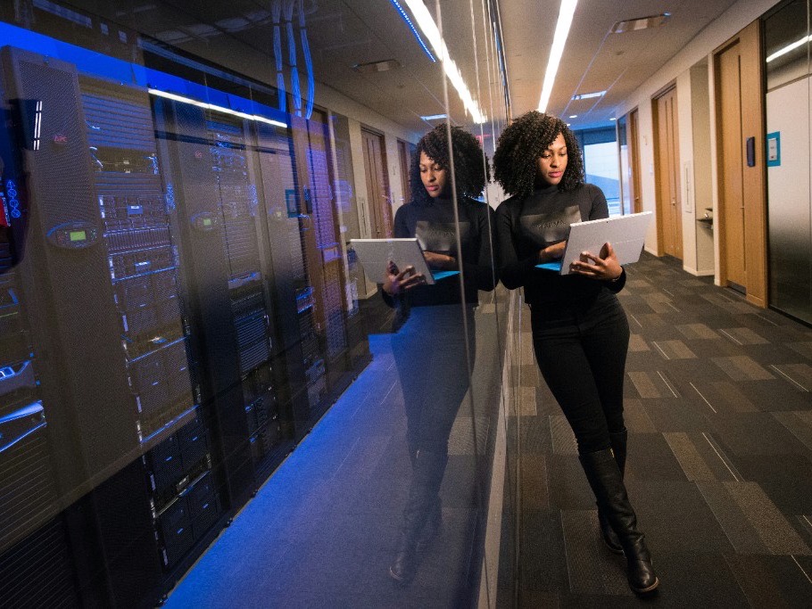 Engineer with a laptop beside a glass-walled server room, representing continuous infrastructure assurance.