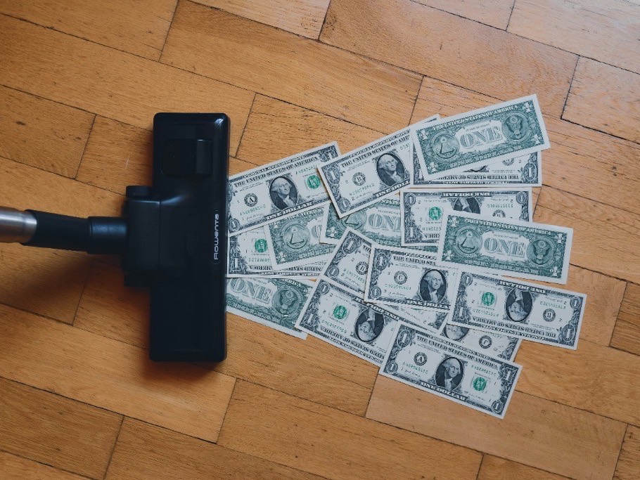 Vacuum nozzle gathering dollar bills on a wooden floor, illustrating cloud cost waste being removed without cutting capacity.